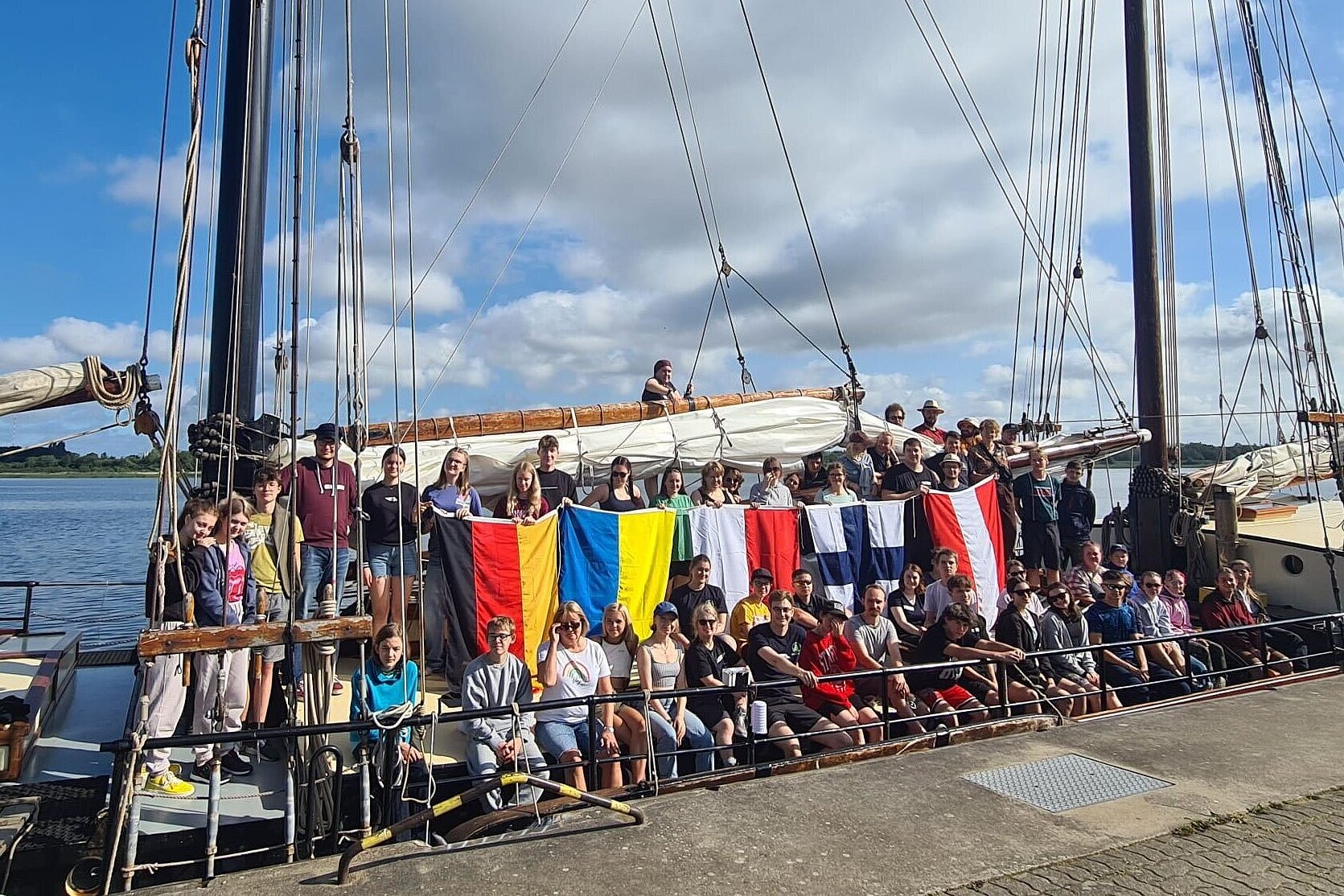 Gruppenfoto, ca. 60 Jugendliche stehen mit den Flaggen von Österreich, Finnland, Polen, Ukraine und Deutschland an Bord eines Segelschiffes.
