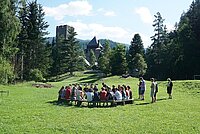 Auf einer grünen Wiese sitzen junge Menschen im Kreis, die Wiese ist von Bäumen links und rechts umgeben, im Hintergrund sind Berge und eine Burg erkennbar.