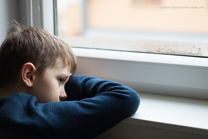 Ein kleiner Junge schaut traurig aus dem Fenster. Seinen Kopf hat er auf die Unterarme auf dem Fensterbrett gelegt.