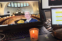 Bildschirm eines Laptops, der den Blick in eine Kapelle zeigt. Im Vordergrund ein Mann mittleren Alters. Auf der Tastatur steht eine brennende Kerze. Daneben ein Bildschirm mit Texten. 