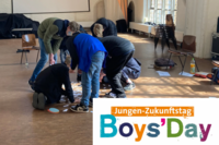 Eine Gruppe Jungen stehen und knien im Kreis in einem großen Saal. Sie gucken auf Papierschnipsel, die auf dem Boden verteilt sind. Unten rechts in der Ecke ist das Logo vom Jungen-Zukunftstag - Boys' Day zu sehen.