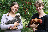 Bild von Svenja Büchner und Marlies Neustadt. Beide haben je ein Huhn im Arm. Im Hintergrund ein Baum mit roten Äpfeln.