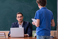 Ein Jugendlicher steht vor dem Schreibtisch eines Lehrer, der von seinem Laptop aufblickt. Im Hintergrund eine Schultafel.