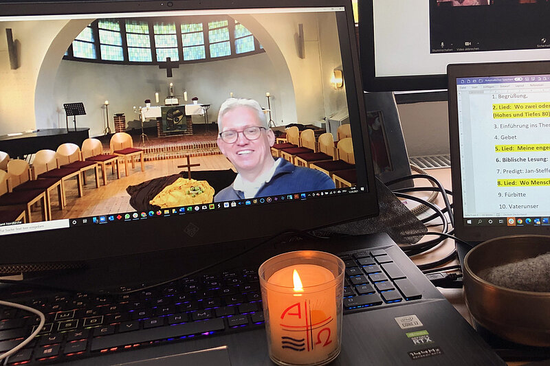 Bildschirm eines Laptops, der den Blick in eine Kapelle zeigt. Im Vordergrund ein Mann mittleren Alters. Auf der Tastatur steht eine brennende Kerze. Daneben ein Bildschirm mit Texten. 