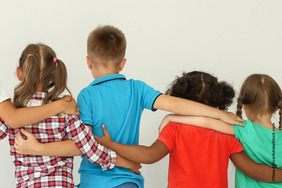 4 Kinder, verschiedener Hautfarbe, in bunter Kleidung, stehen vor einem grauen Hintergrund. Zusehen sind sie von hinten. Sie stehen nebeneinander und haben einander die Arme auf die Schultern gelegt.