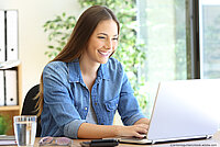 Eine junge Frau am Tisch sitzend, schreibt mit freudigem Gesichtsausdruck auf ihrem Laptop.