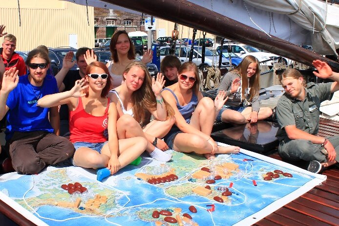 Eine Gruppe junger Menschen sitzt an Bord eines Schiffes auf einer großen Weltkarte und winkt fröhlich.