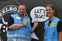 Ein Mann und eine Frau in blauen Westen mit der gelben Aufschrift "Seelsorge" Im Hintergrund der Schriftzug: "Seelsorge" sowie "Let´s talk".