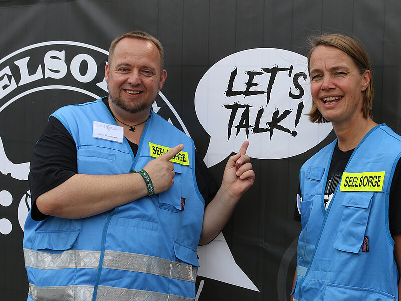 Ein Mann und eine Frau in blauen Westen mit der gelben Aufschrift "Seelsorge" Im Hintergrund der Schriftzug: "Seelsorge" sowie "Let´s talk".
