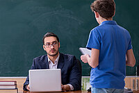 Ein Schüler steht vor dem Lehrerschreibtisch. Der Lehrer blickt von seinem Laptop auf. Im Hintergrund eine grüne Schultafel.