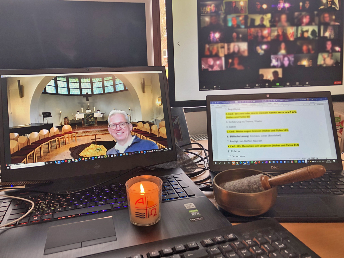 Bildschirm eines Laptops, der den Blick in eine Kapelle zeigt. Im Vordergrund ein Mann mittleren Alters. Auf der Tastatur steht eine brennende Kerze. Daneben ein Bildschirm mit Texten. Daneben ein Bildschirm mit "Zoom-Kacheln".